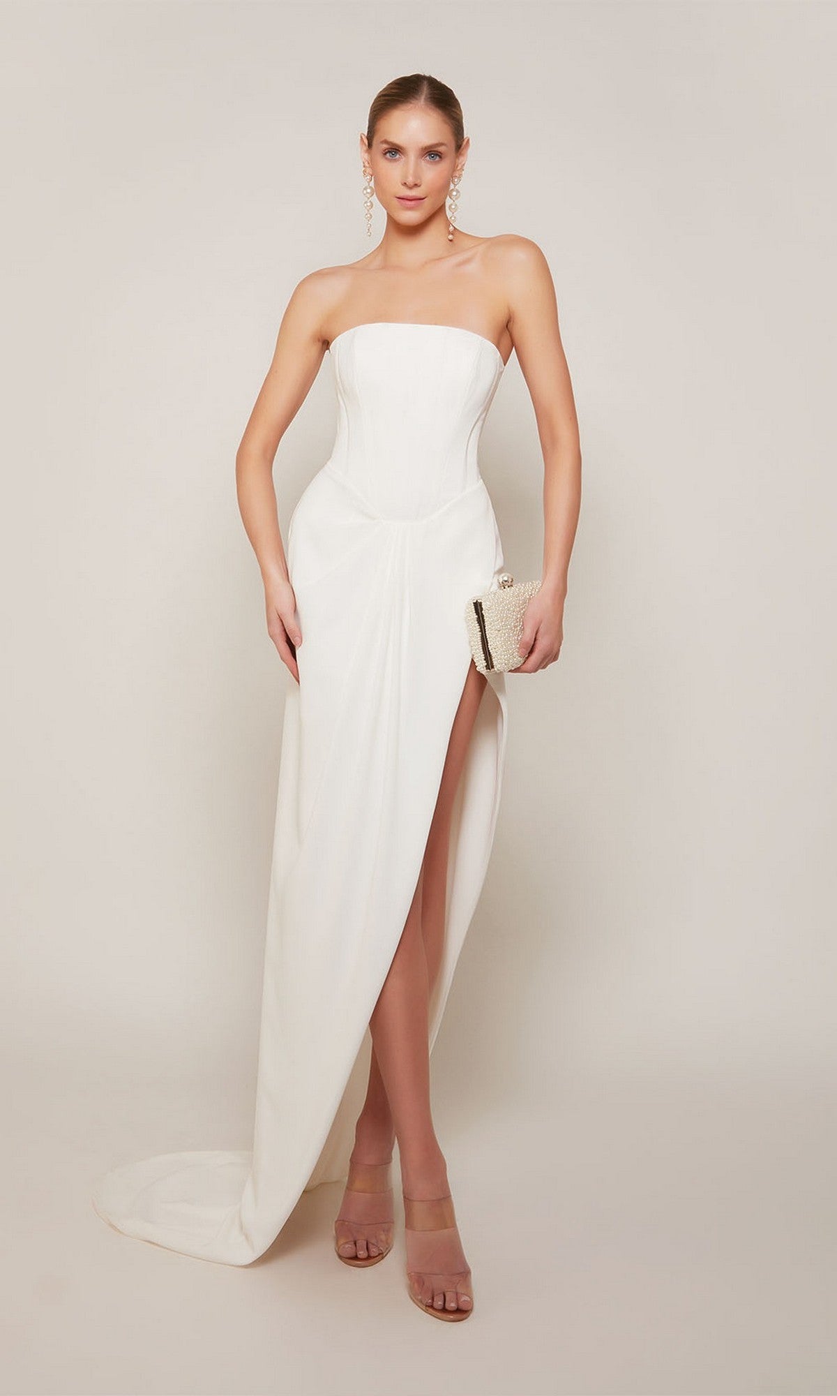 A woman in the Alyce 70042 long formal dress—strapless, white, with a corset bodice and thigh-high slit—stands against a plain background holding a clutch, wearing nude heels and drop earrings.